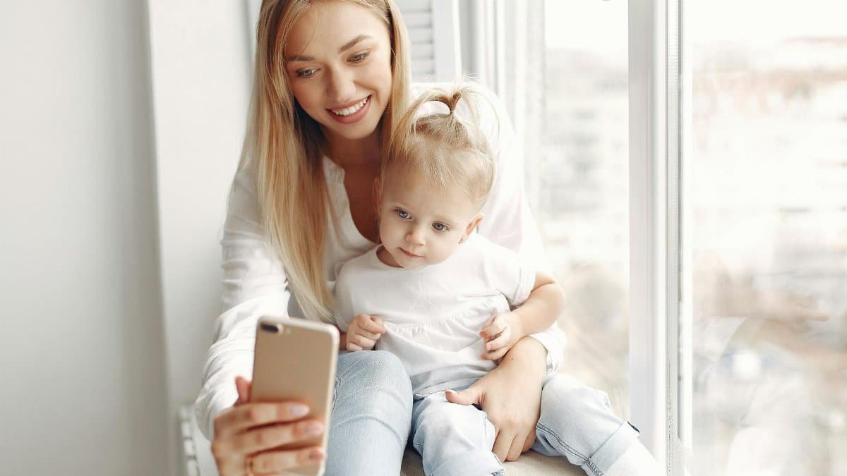 A mother and daughter bonding indoors by a window, sharing a moment with a smartphone.