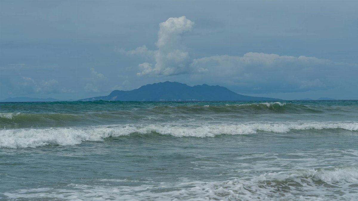 Serene beach view with gentle waves and a distant island under a cloudy sky in New Zealand.