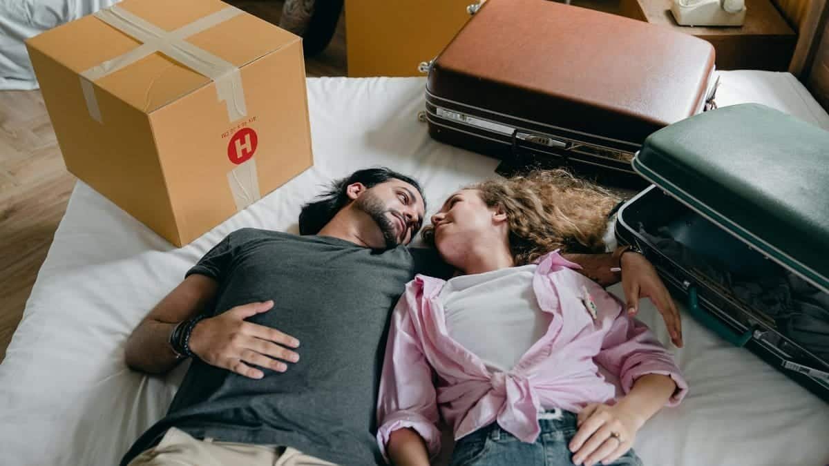 From above of young ethnic bearded man and woman with curly hair looking at each other and lying on bed among suitcases and cardboard boxes with stuff while moving in new apartment together