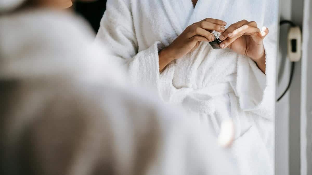 Crop unrecognizable young lady in white bathrobe opening face cream while standing in front of mirror after taking shower in morning