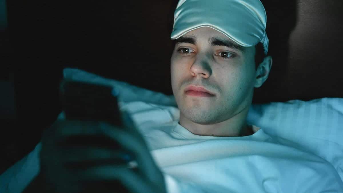 A man wearing a sleeping mask uses his smartphone while lying in bed at night.