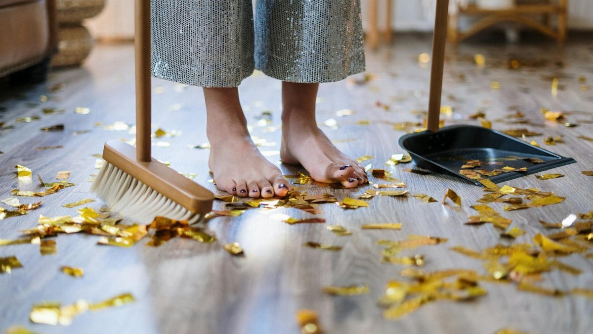 Barefoot woman sweeping gold confetti with broom and dustpan indoors.