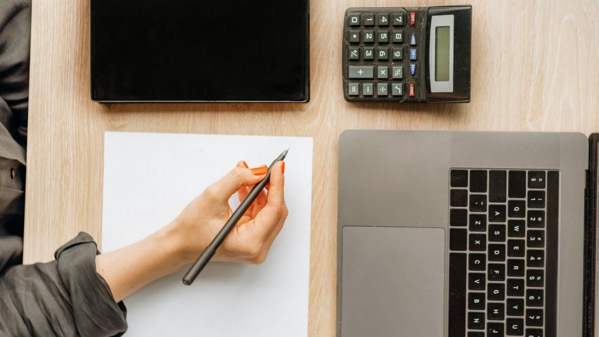 A person writing on paper surrounded by a laptop, tablet, and calculator on an office desk.
