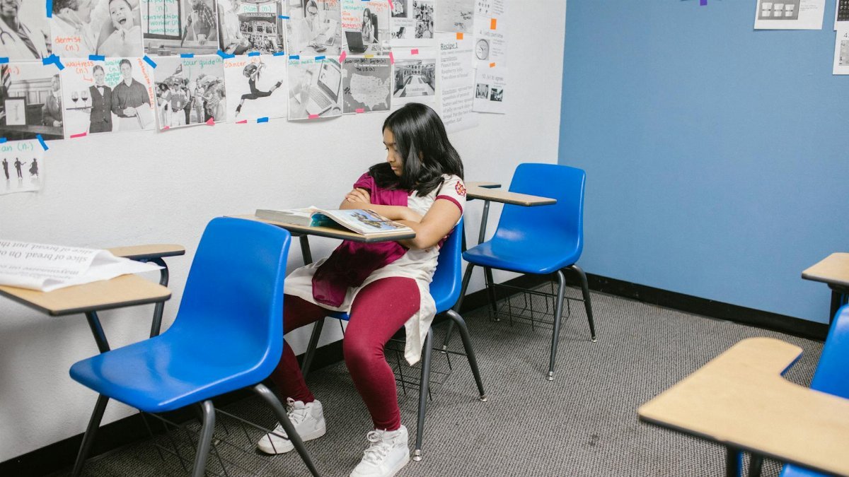 Young girl sitting alone in a classroom with blue chairs, symbolizing bullying and isolation.