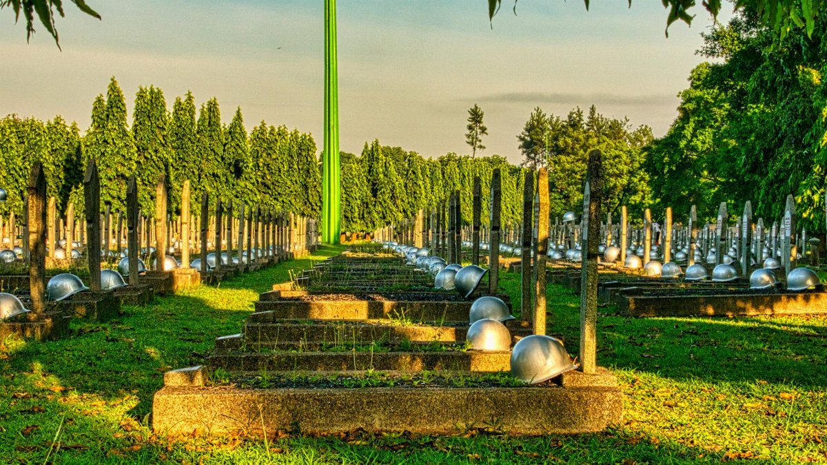 Various tombstones with military hardhats located on grassy ground in national main heroes cemetery against green trees and tall structure