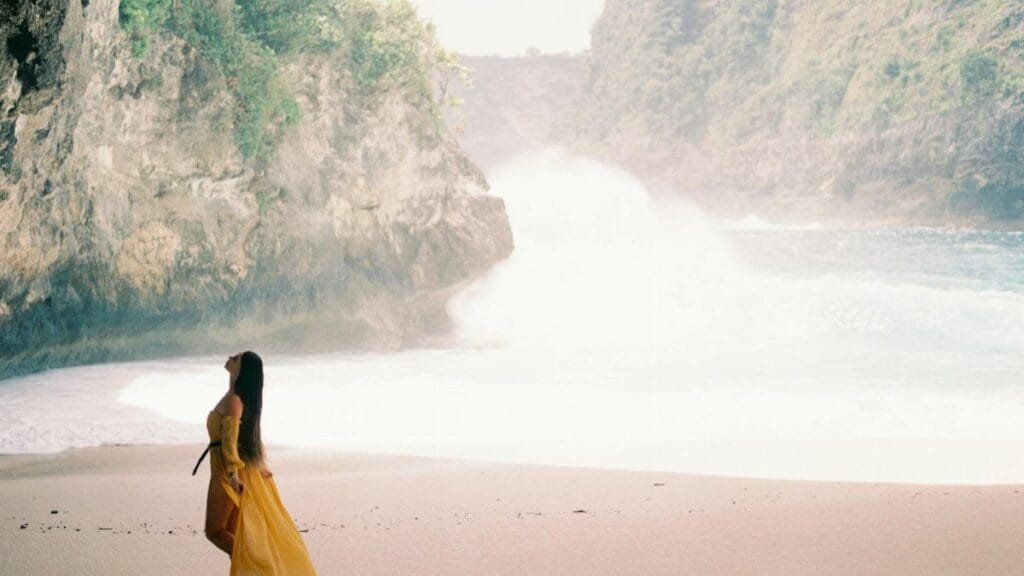 A woman in a flowing yellow dress walking along a peaceful beach with cliffs and waves.