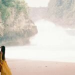 A woman in a flowing yellow dress walking along a peaceful beach with cliffs and waves.