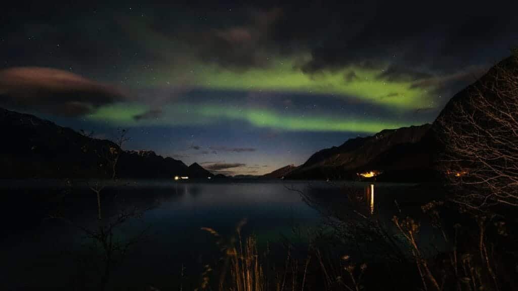 Beautiful view of the northern lights reflecting over a tranquil lake surrounded by mountains at night.