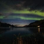 Beautiful view of the northern lights reflecting over a tranquil lake surrounded by mountains at night.