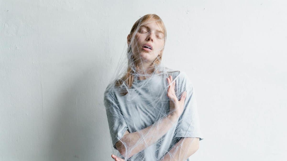 A woman wrapped in a spider web against a white wall, expressing feelings of entrapment and anxiety.