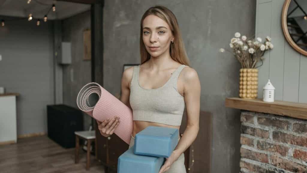 A woman in a sports bra holds yoga mat and blocks, standing in a modern studio setting.