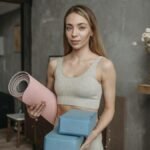 A woman in a sports bra holds yoga mat and blocks, standing in a modern studio setting.