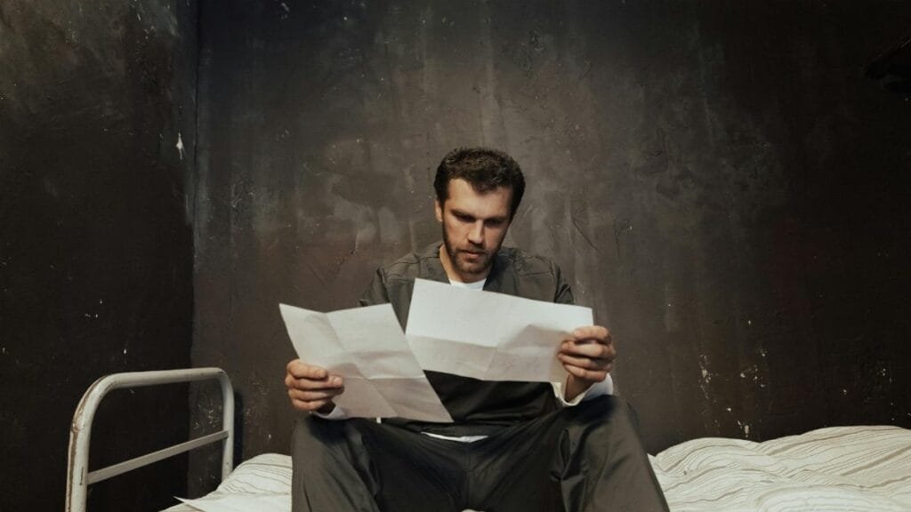 A prisoner sits on a bed in a cell reading papers, highlighting confinement themes.
