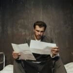A prisoner sits on a bed in a cell reading papers, highlighting confinement themes.