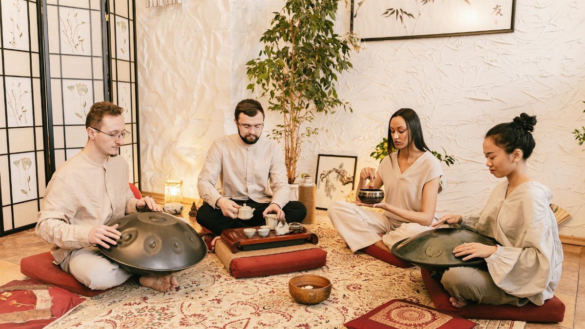 A diverse group meditates with handpans and tea in a serene, indoor setting.