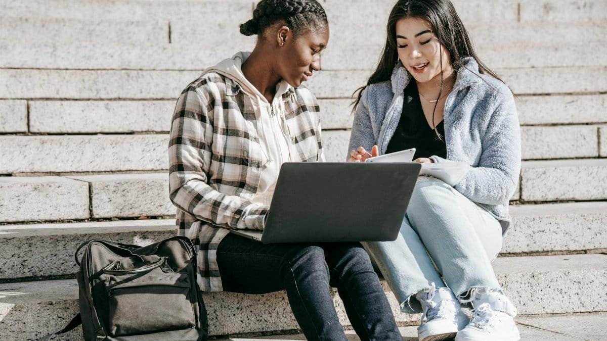 Two diverse students collaborating on laptops and notepads outdoors on university steps.