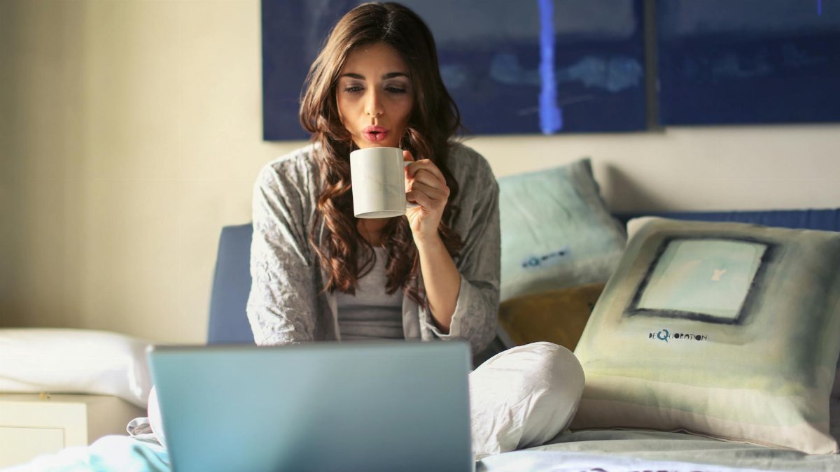A woman enjoying coffee while working from home in a cozy bedroom setting with a laptop.