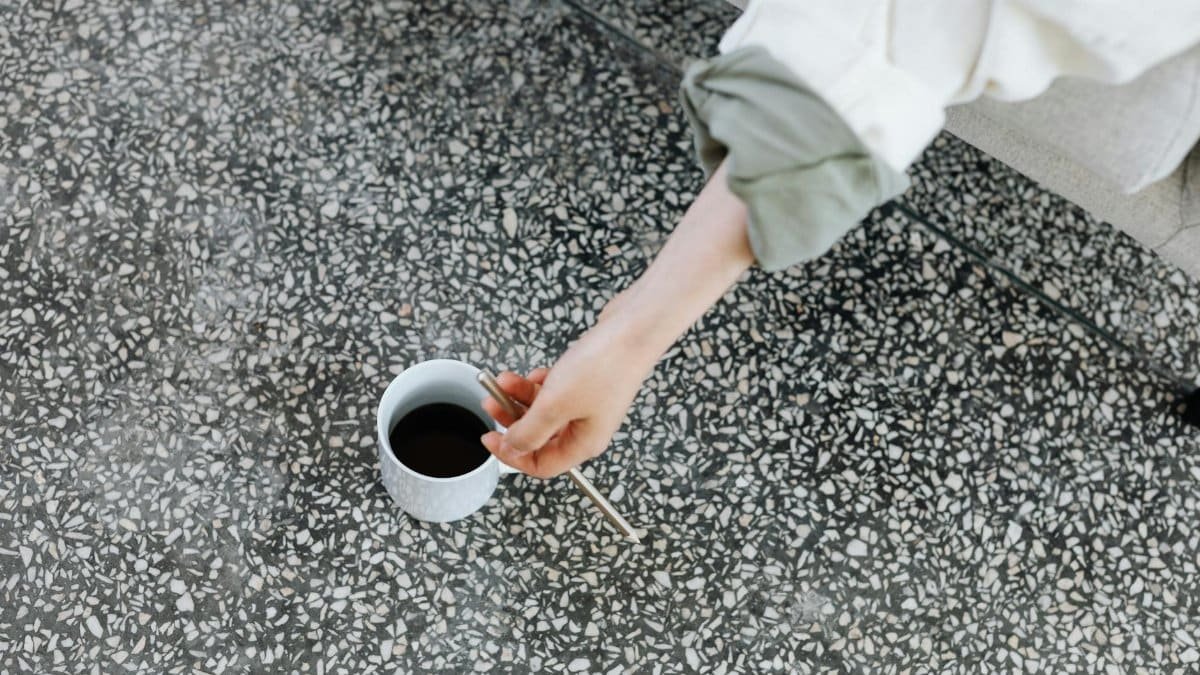 A person reaches for a cup of coffee placed on a terrazzo floor, creating an everyday casual scene.