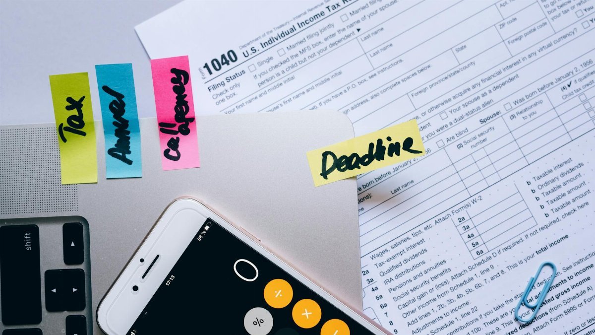 Office desk flat lay showing tax documents, calculator app on smartphone, sticky notes, and paperclips.
