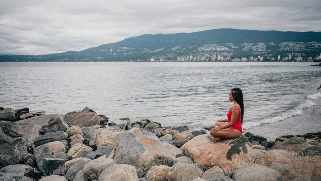 A woman in a red swimsuit meditates on a rocky shore by the ocean under a cloudy sky.