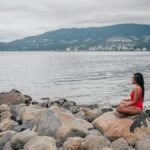 A woman in a red swimsuit meditates on a rocky shore by the ocean under a cloudy sky.