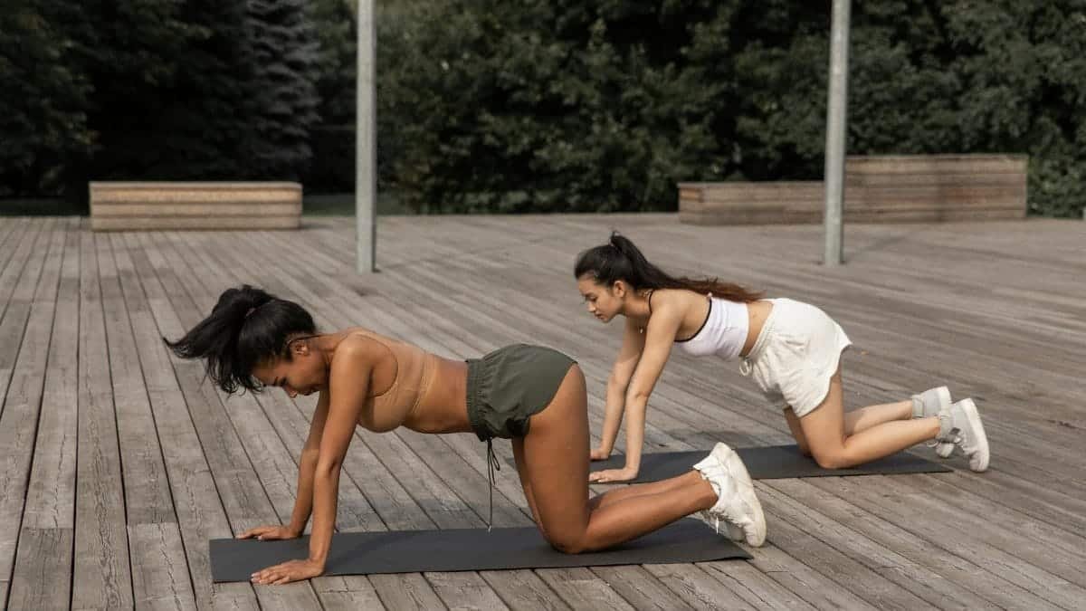 Two women in active wear practicing yoga on a wooden deck outdoors, promoting wellness and mindfulness.