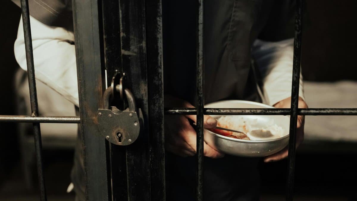 Close-up of a prisoner holding a bowl behind iron bars, illustrating confinement.