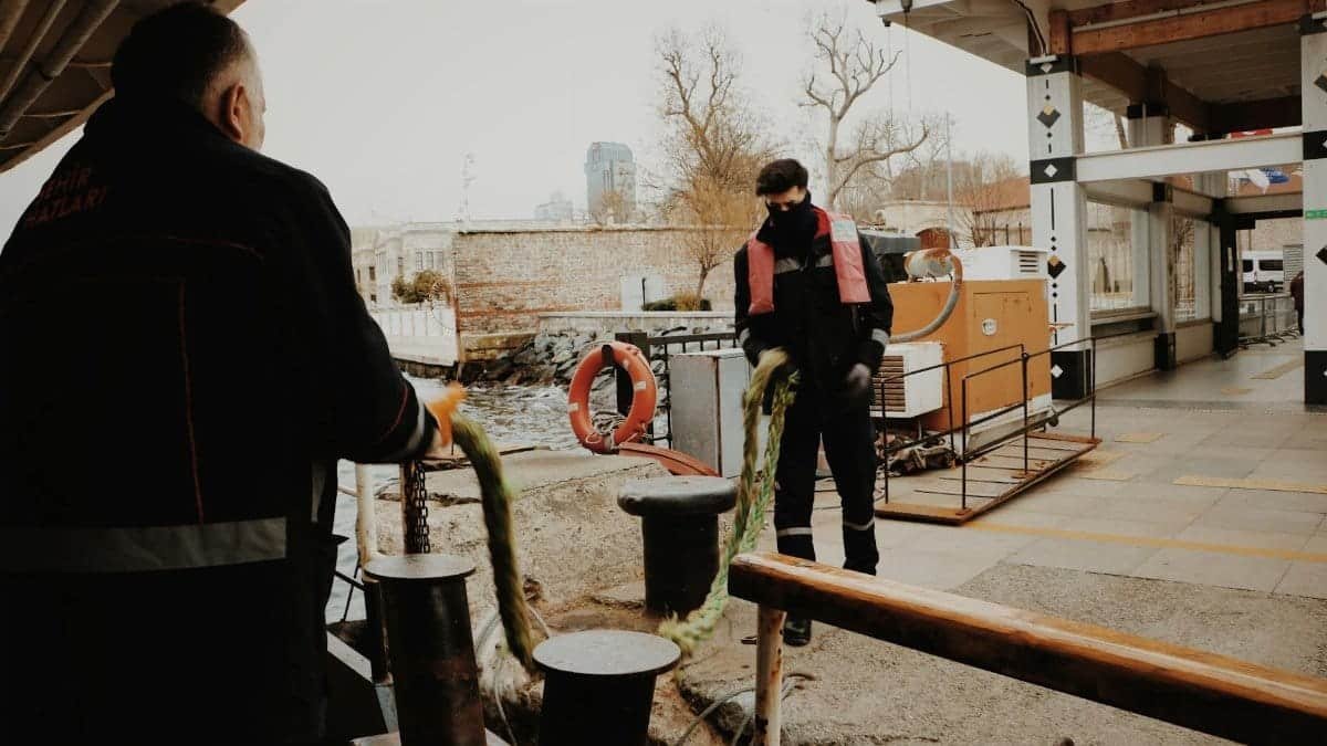 Urban scene of dock workers managing ropes at a port with buildings in the background.
