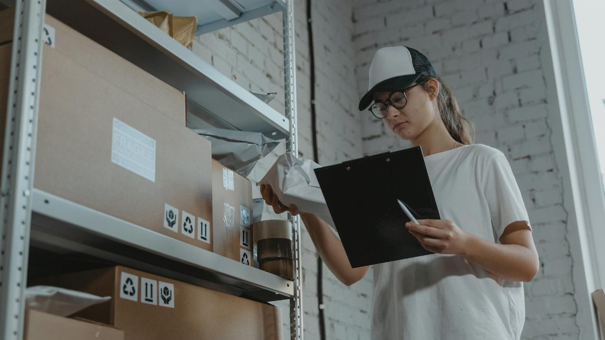 Warehouse worker organizing packages on shelf with clipboard.