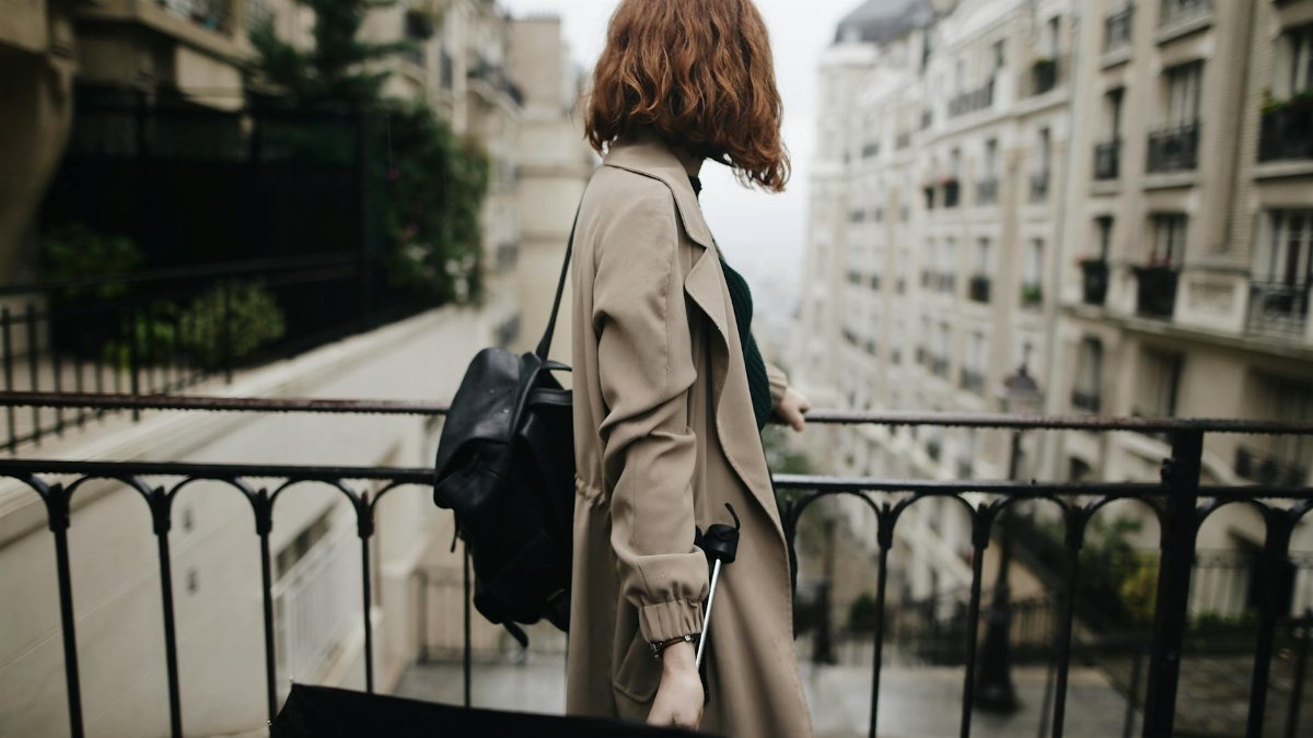 Woman in trench coat walking through a Parisian street with an umbrella, showcasing city life and travel.