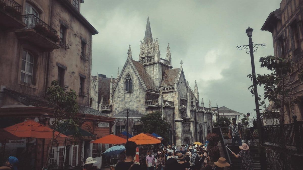 A gothic revival church in a busy urban setting with tourists and vibrant umbrellas on a cloudy day.