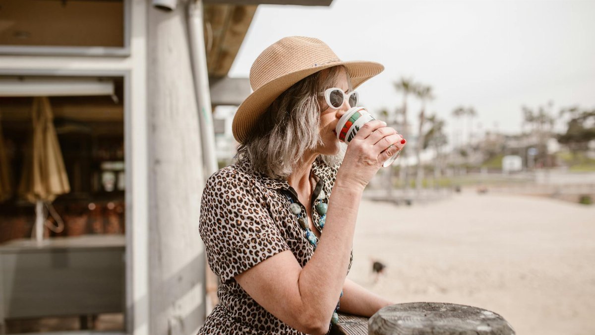Stylish woman in leopard print and hat enjoying coffee outside a cafe by the beach.