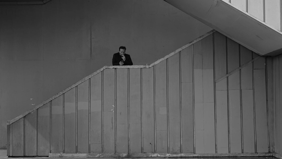 Black and white emotionless adult male in black jacket smoking cigarette and standing on outdoor building stairway