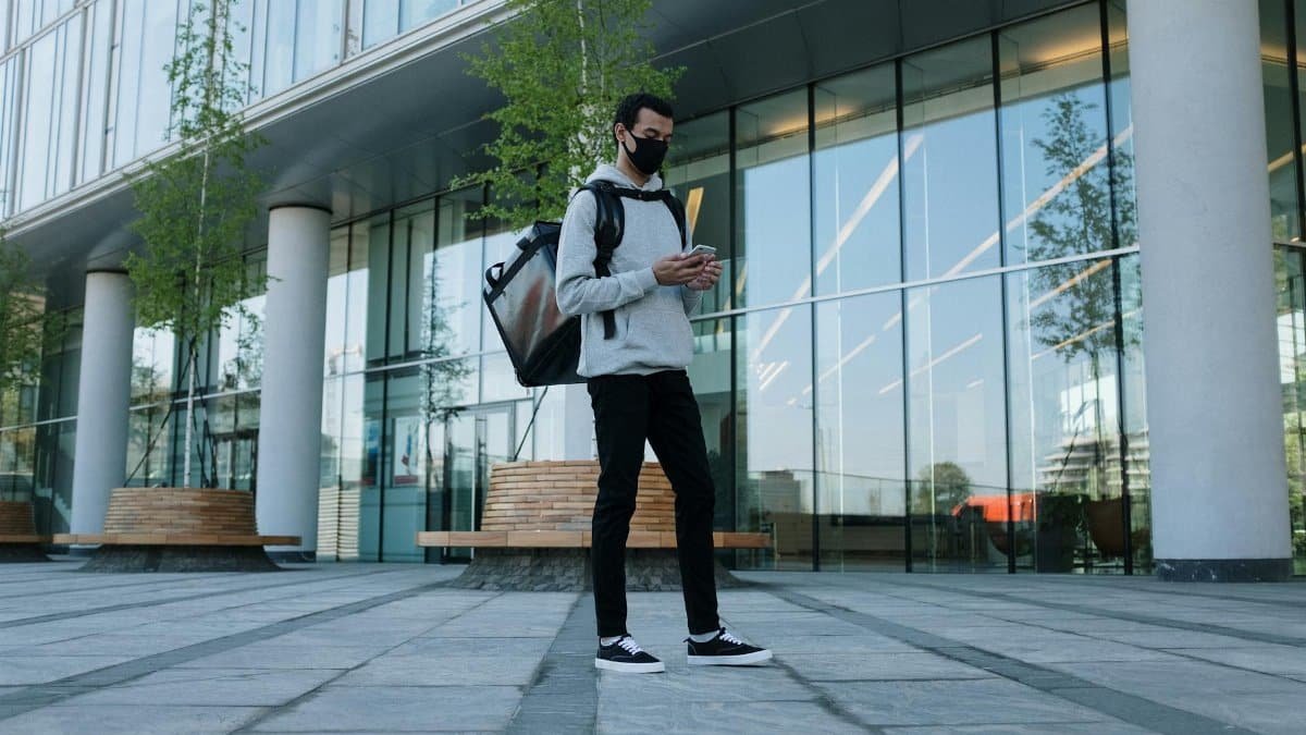A masked courier in a city setting delivering a package amidst modern architecture.