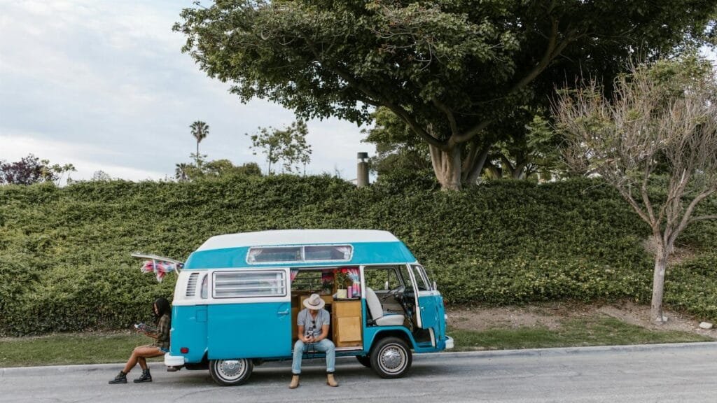 A couple enjoying a sunny day next to a retro blue camper van parked by greenery.