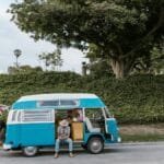 A couple enjoying a sunny day next to a retro blue camper van parked by greenery.