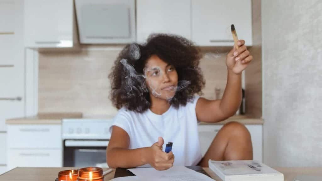 A woman in a kitchen using a smudge stick for cleansing, surrounded by papers and candles.