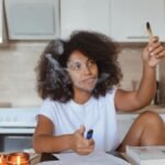 A woman in a kitchen using a smudge stick for cleansing, surrounded by papers and candles.