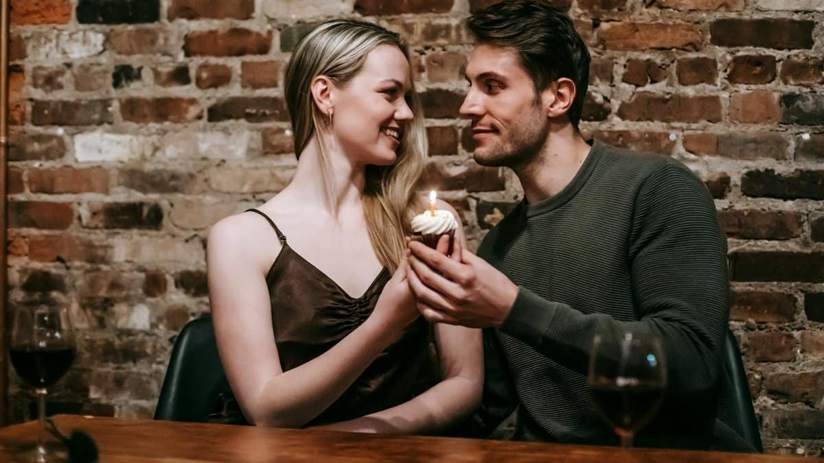 Happy young couple in elegant clothes sitting at table and celebrating birthday with small cupcake and candle near glasses with red wine and brick wall while looking at each other