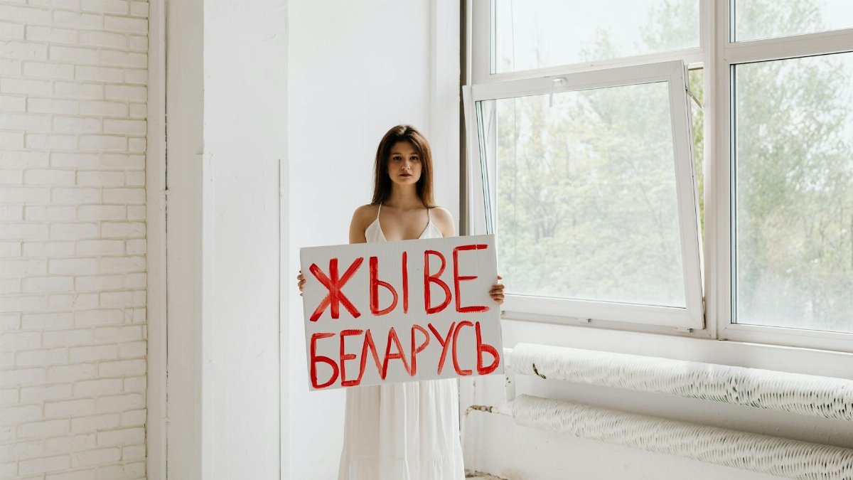 A woman stands indoors holding a sign with red text in a peaceful protest setting.