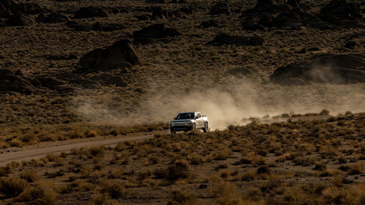 A white truck kicks up dust on a rugged off-road desert trail surrounded by rocky terrain.