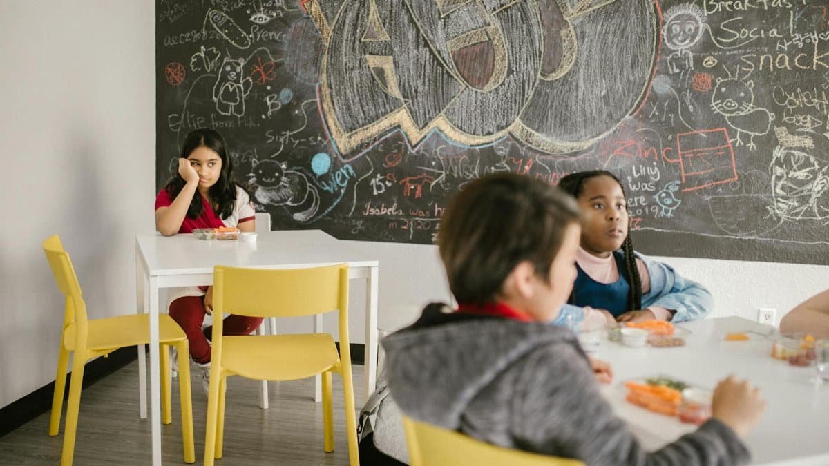 Children sitting in classroom, one girl alone, with chalkboard featuring graffiti artwork and text.