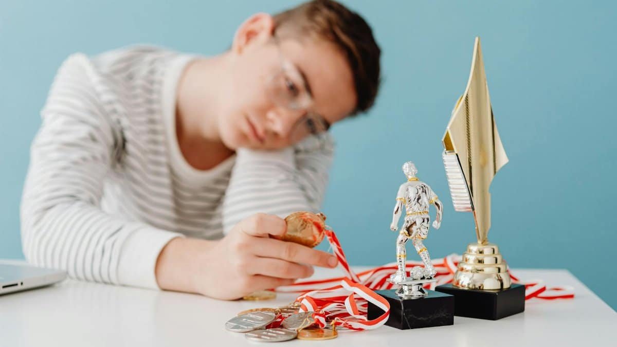 A thoughtful teenager sits by a table with various trophies and medals.