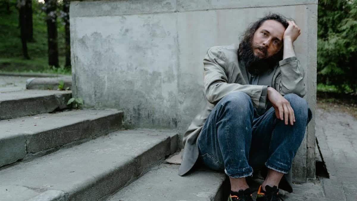 A homeless man sitting on stone steps showing signs of sadness and fatigue.