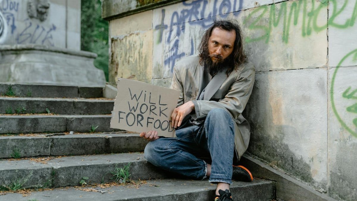 A homeless man sits on steps outdoors holding a 'Will Work for Food' sign, depicting poverty.
