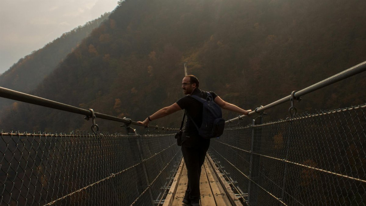 A man with a backpack crossing a suspension bridge in a foggy mountain setting.
