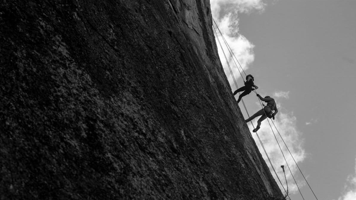 A dramatic black and white shot of two climbers scaling a sheer rock face against a cloudy sky.