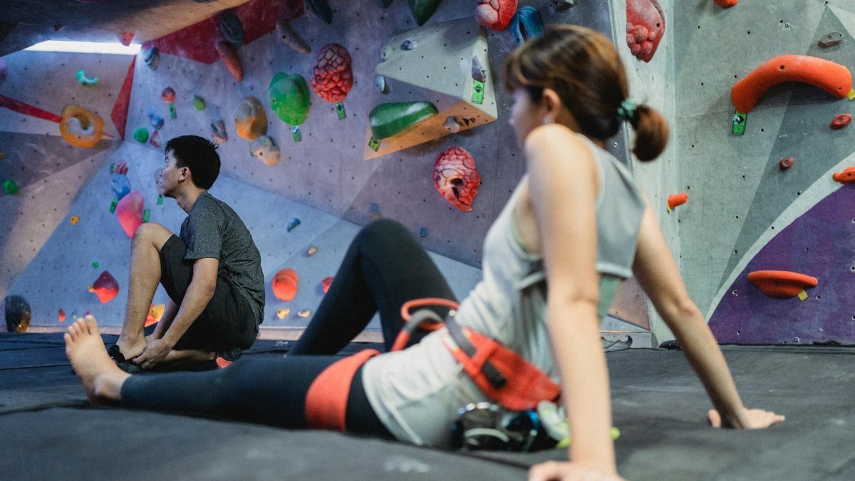 Two young adults resting on mats in an indoor bouldering gym, surrounded by colorful climbing holds.