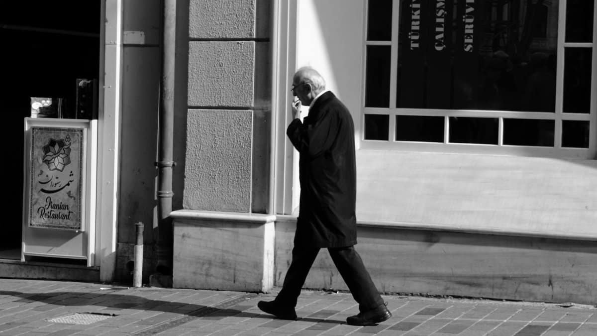 Elderly man walking while smoking on a city sidewalk next to a building window.