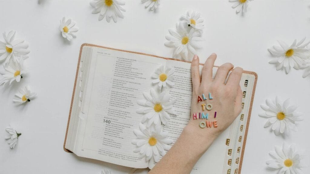 Open Bible adorned with daisies and letters spelling 'ALL TO HIM I OWE', emphasizing faith and inspiration.